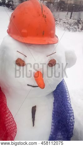 A Snowman In An Orange Helmet With Walnut Shell Eyes And A Mesh Scarf. An Orange Carrot Like The Nos