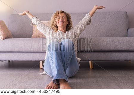 Happy And Satisfied Woman At Home Sitting On The Floor Opening Arms And Smiling. Concept Of Joyful L