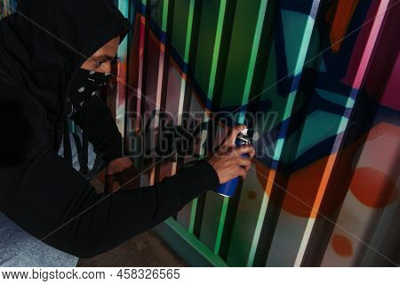 Side View Of African Amercan Hooligan In Mask Drawing Graffiti On Wall At Night