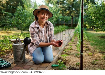 Caucasian Pretty Woman, Farmer Sitting Near A Watering Can, Holding A Seedling Of Cucumbers, Smiling