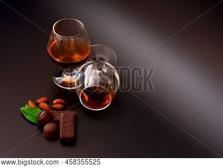 Two Glasses Of Cognac, Chocolates, Almond Kernels And Mint Leaves On A Black Background.