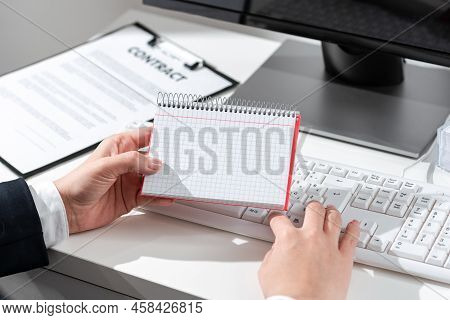 Businesswoman Holding Important Message Wtitten On Notebook On Desk With Computer, Clipboard And Not