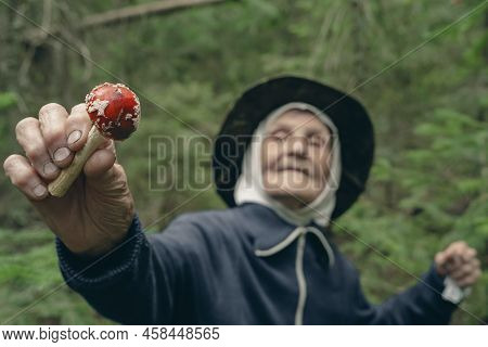 An Elderly Woman Holds A Red Fly Agaric In Her Hands Against The Background Of The Forest. The Old L