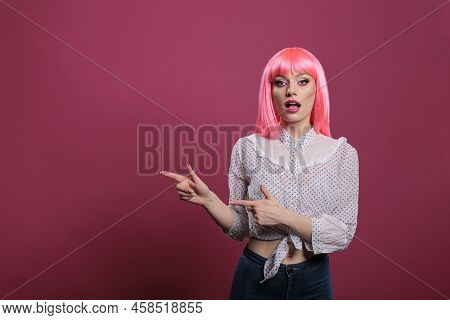 Attractive Young Woman With Pink Hair Pointing Sideways In Studio, Indicating Left Or Right Directio