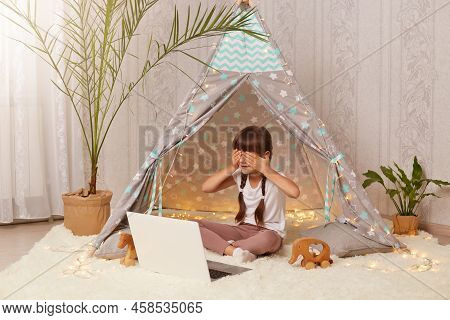 Indoor Shot Of Little Preschooler Girl Sitting In Wigwam In Front Of Notebook, Having Video Call, Co