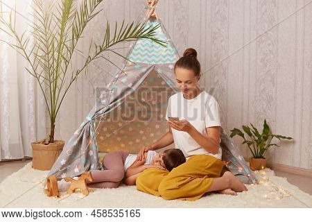Indoor Shot Of Happy Caucasian Young Adult Woman Sitting On Floor Near Children's Wigwam And Using C