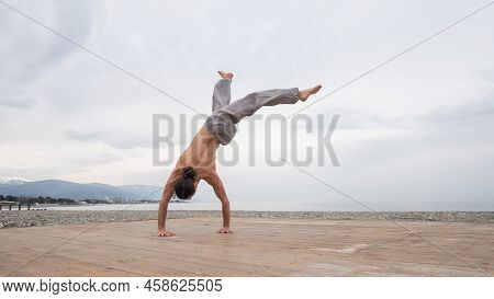 Shirtless Caucasian Man Doing Backflip On Pebble Beach.