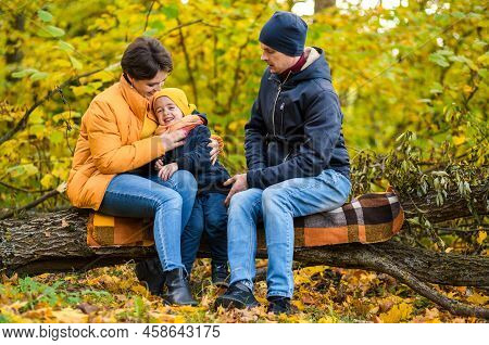 Mother, Father And Little Son Sit On A Plaid On A Log In The Autumn Park And Spend Time Together. Ho