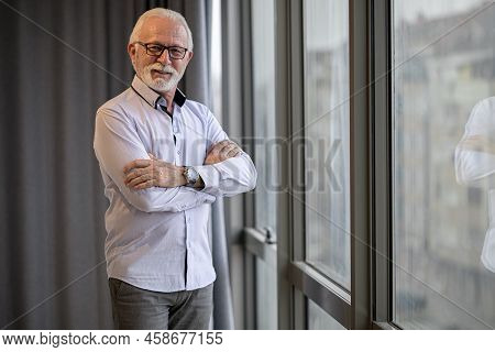Portrait Of Determined Senior Entrepreneur By Window. Confident Elderly Businessman Is Standing With