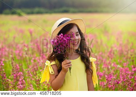 A Beautiful Playful Little Girl In A Straw Hat, Holds A Bouquet Of Burgundy Viscaria Flowers