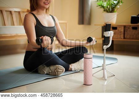 Smiling Young Woman Following Tutorial On Smartphone When Doing Exercise With Dumbbells