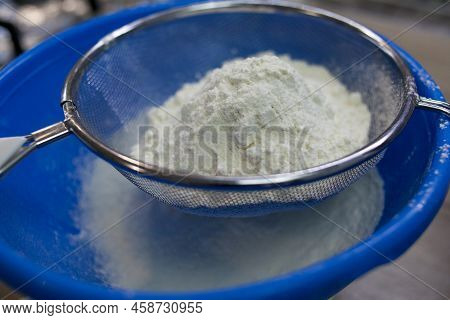 Sifting White Flour Through A Sieve Into Blue Bowl On Table On Wooden Background. Flour For Dough Pr