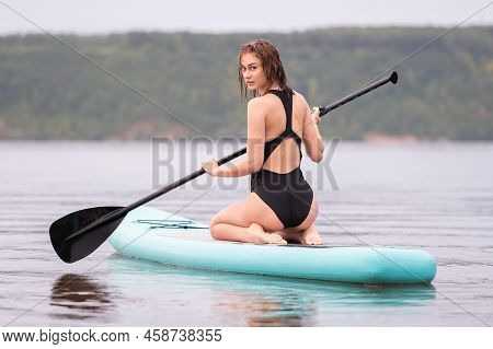 A Beautiful Girl Is Floating On A Supboard On A River. Young Woman Supsurfing At Sunrise.