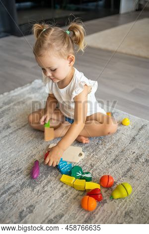 Cute Little Toddler Girl Playing At Home With Eco Wooden Toys. Happy Child Cutting Vegetables And Fr