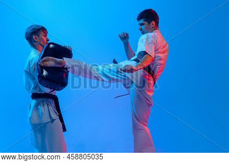 Studio Shot Of Sports Training Of Two Karatedo Fighters In Doboks Isolated On Blue Background In Neo