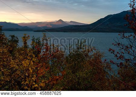Sun Rises In The Fog, Making Sky Yellow, And Illuminates Dramatic Mountain Peaks Above Lake In Remot