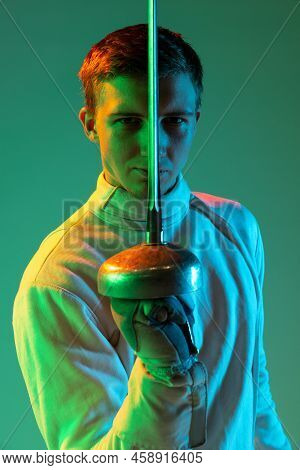 Portrait Of Young Tall Male Fencer In Fencing Costume And Mask Holding Smallsword Isolated On Green 