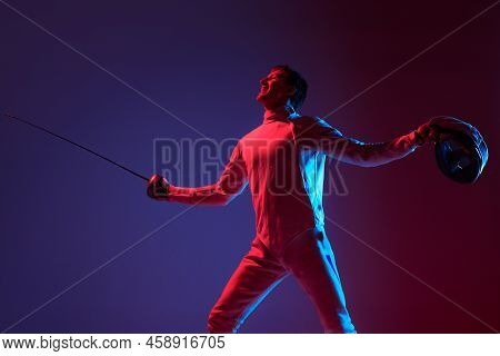 Portrait Of Young Tall Male Fencer In Fencing Costume And Mask Holding Smallsword Isolated On Purple