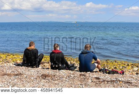 Three People Sit On The Seashore In Diving Clothes And Look Into The Distance.