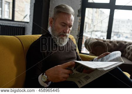 Senior Focused Caucasian Man Reading Newspaper On Sofa At Home. Stylish Bearded Pensioner Wearing Ca