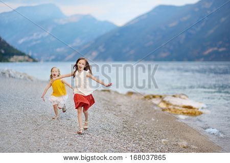 Two Cute Little Sisters Having Fun On A Beach Of Limone Sul Garda, A Small Town And Comune In The Pr