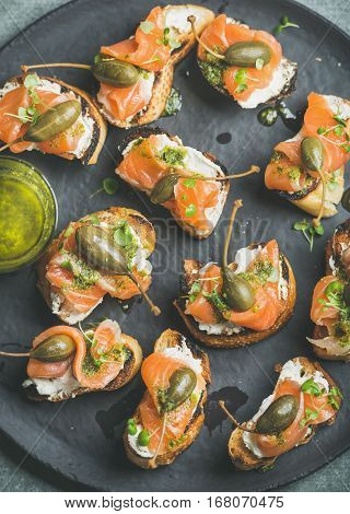 Homemade salmon crostini with cream-cheese, watercress, capers and pesto suace in dark slate stone plate, top view, vertical composition