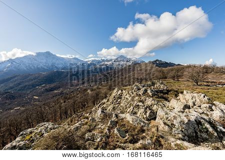 View of snow covered San Parteo viewed from Col de Battaglia in the Balagne region of Corsica with a rocky outcrop in the foreground and blue skies above