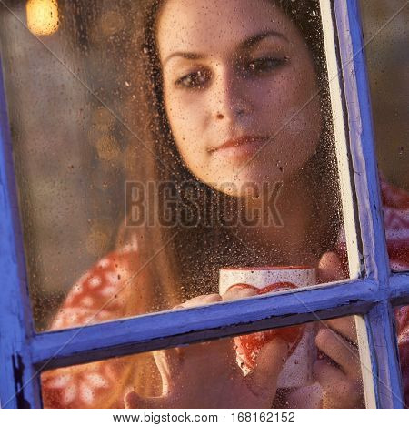Pretty woman in warm room indoor at home in winter night. Drinking tea looking away. Candid shot from outdoor trough the window.
