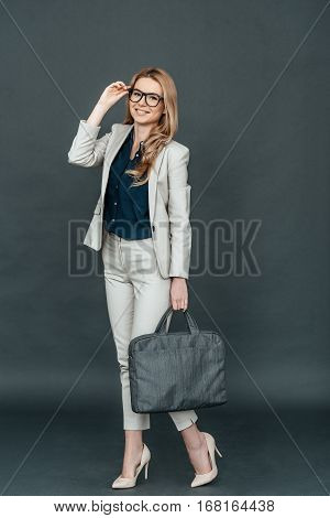 Young and smart. Full length of confident young woman in smart casual wear adjusting her eyeglasses and looking at camera while standing against grey background