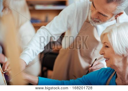 Useful cooperation. Professional male artist helping his elderly female colleague while drawing picture and spending time in painting class together.