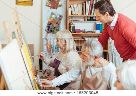 Talented teacher. Concentrated handsome young artist controlling his colleagues while sitting in painting class and drawing together