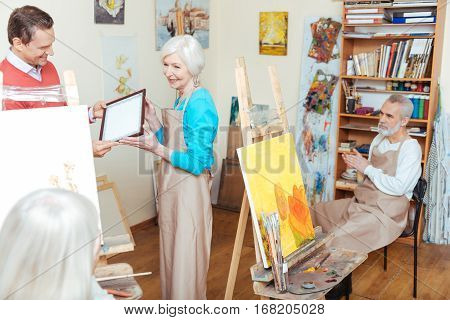 Be proud. Handsome young smiling man giving artists diploma to woman while spending time in painting school and working as a teacher.