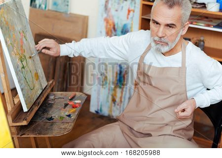 Inspiration in calmness. Concentrated elderly professional artist painting a picture while working and sitting in painting class.