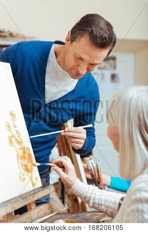 Good results. Serious handsome concentrated man teaching elderly woman while drawing pictures in painting school and enjoying their hobby.