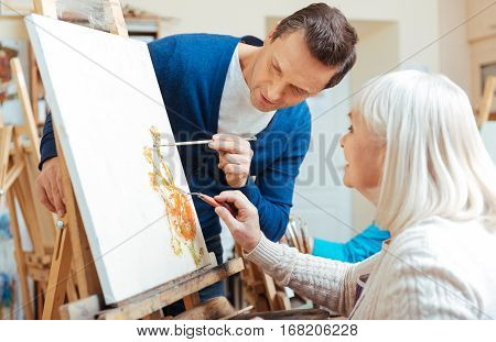 Perfect result. Serious handsome concentrated man teaching elderly woman while drawing pictures in painting school and enjoying their hobby.