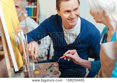 Best teacher. Friendly handsome young artist helping elderly woman while she painting and spending time in painting studio
