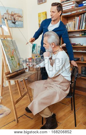 Perfect worker. Ambitious handsome young artist helping elderly man while he painting and spending time in painting studio