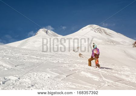 Stylishly dressed snowboarder stands and walks on mountain slopes of an extinct volcano Elbrus on a background of pure blue sky