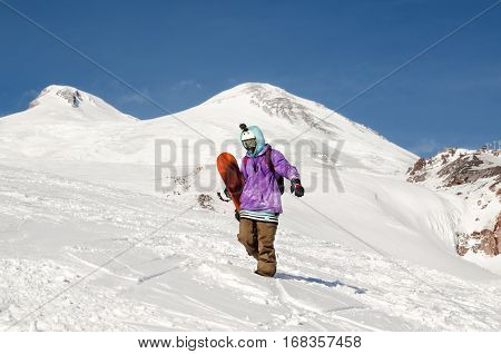 Stylishly dressed snowboarder stands and walks on mountain slopes of an extinct volcano Elbrus on a background of pure blue sky