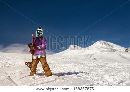 Stylishly dressed snowboarder stands and walks on mountain slopes of an extinct volcano Elbrus on a background of pure blue sky