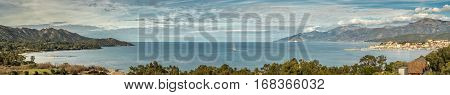 Panoramic view across the bay of Saint Florent in Corsica with rocks in the foreground and a three mast sailing ship in the harbour with the mountains of Cap Corse behind the town of Saint Florent