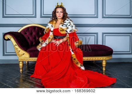 Woman in red dress, cloak and with crown on head sits on couch in room.
