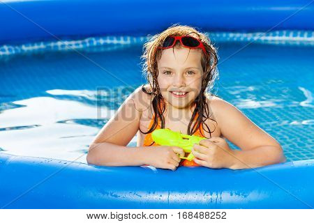 Portrait of happy girl in sunglasses playing with water gun in the blue inflatable pool