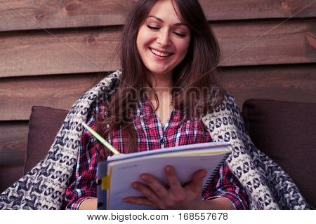 Low angle shot of smiling woman sitting on the sofa, covered with a warm blanket enjoying evening at the home. Good-looking young woman smiling while making notes in a jotter