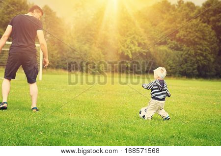 Little boy playting football with father on the field with gates. Active man and child in summer with ball