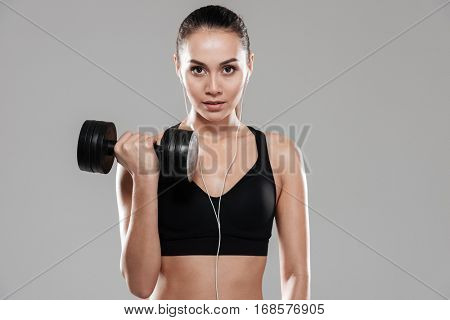 Front view of Sporting woman using dumbbell in studio and looking at camera. Isolated gray background