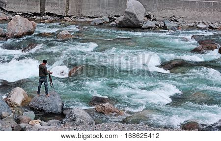 Aadventurous Photographer on dangerous flooded river in Azad Kashmir. standing on a rock and capturing the moment with his camera on tripod.
