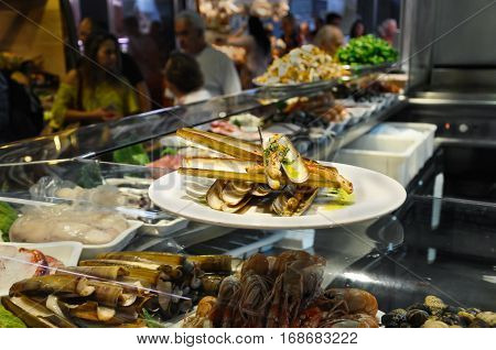 Shrimps bamboo jack knife clams and mussels on ice in glass case of cafe at Mercat de Sant Josep Boqueria market Barcelona Spain. Fried Gould's razor shell or Canyut navaja in olive oil and pesto