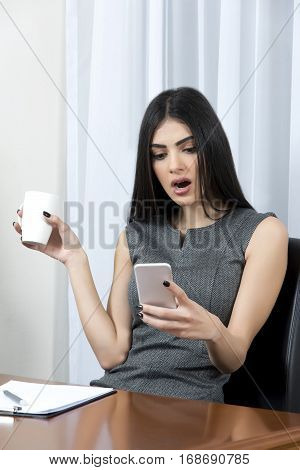 Young business woman sitting at her office at work. In one hand is holding a cup of coffee and at the other her mobile phone.