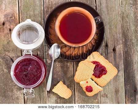 Tea drinking with red berries jam and cookies flat lay. The ceramic cup with tea costs on a table from old boards the jar of jam and pieces of cookies smeared with jam nearby